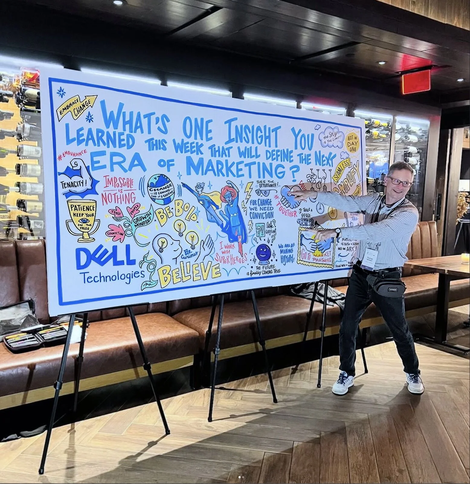A man in glasses and a striped shirt is pointing at a large colorful illustrated board on an easel, inside a restaurant or café decorated with wine bottles. The board features doodles, text, and symbols related to marketing, technology, and inspirati