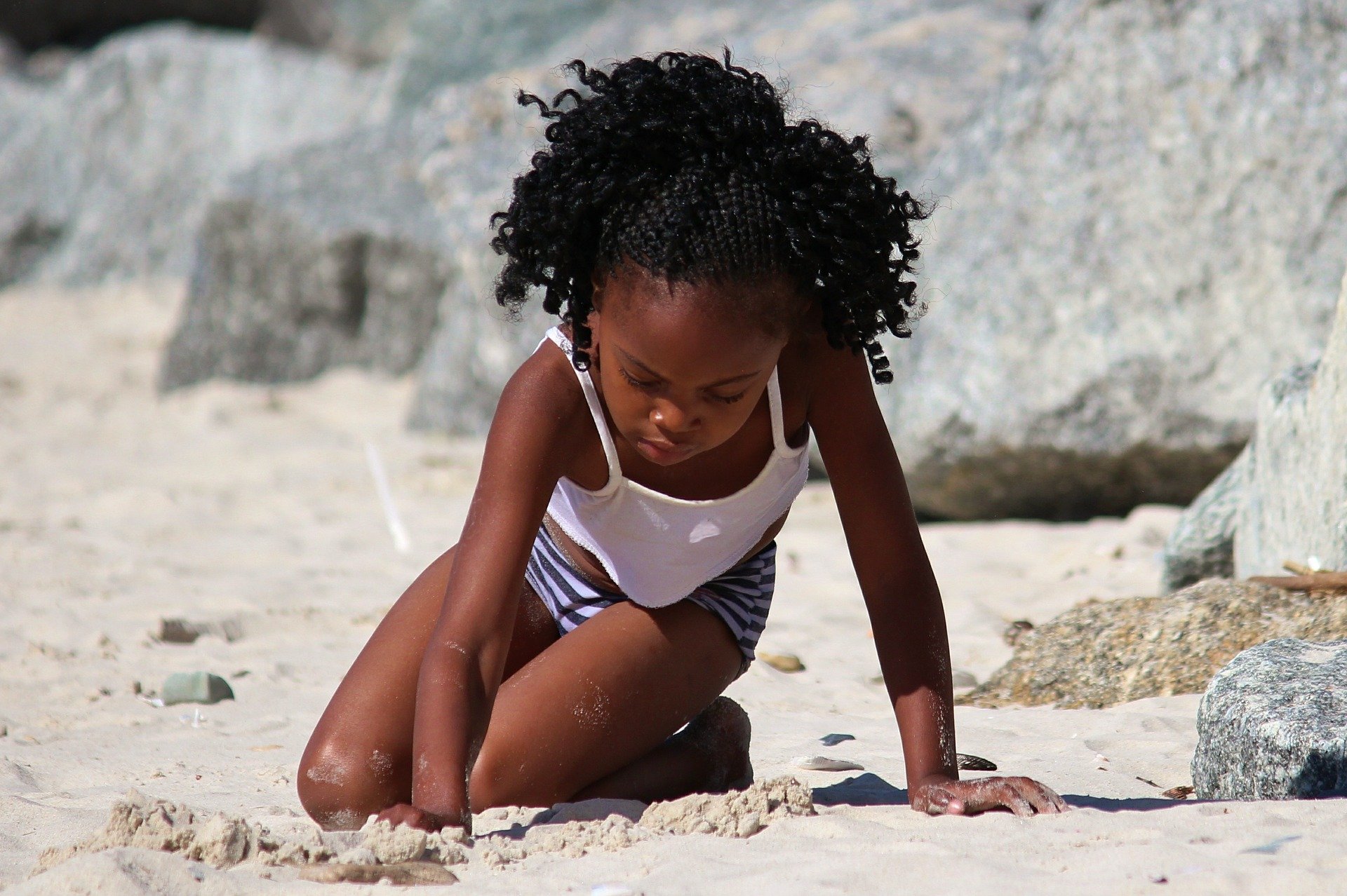 Young girl sitting on a beach with her hand moving the sand representing sensory play.
