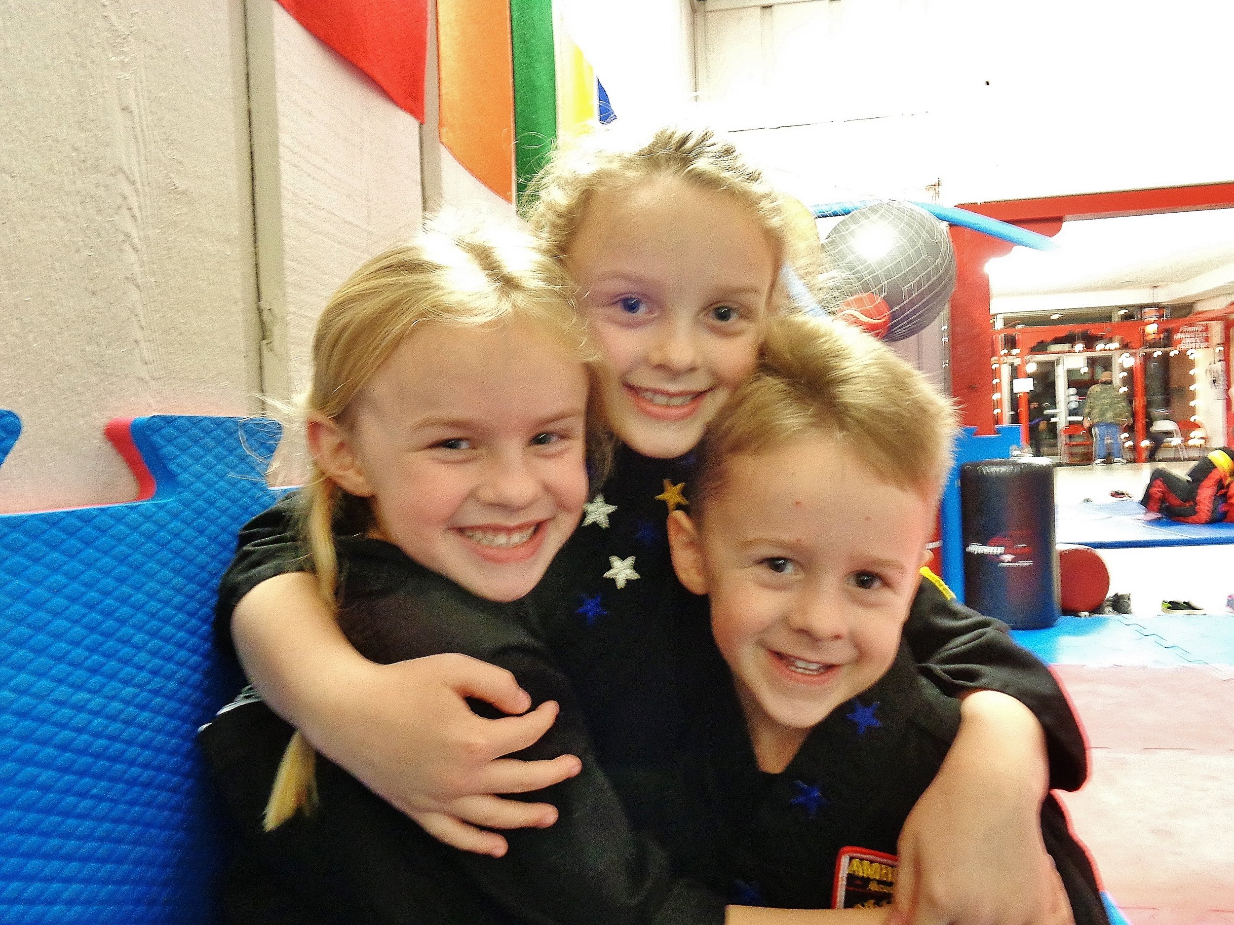 Three martial art kids, two girls, one boy, hugging and smiling at the camera.
