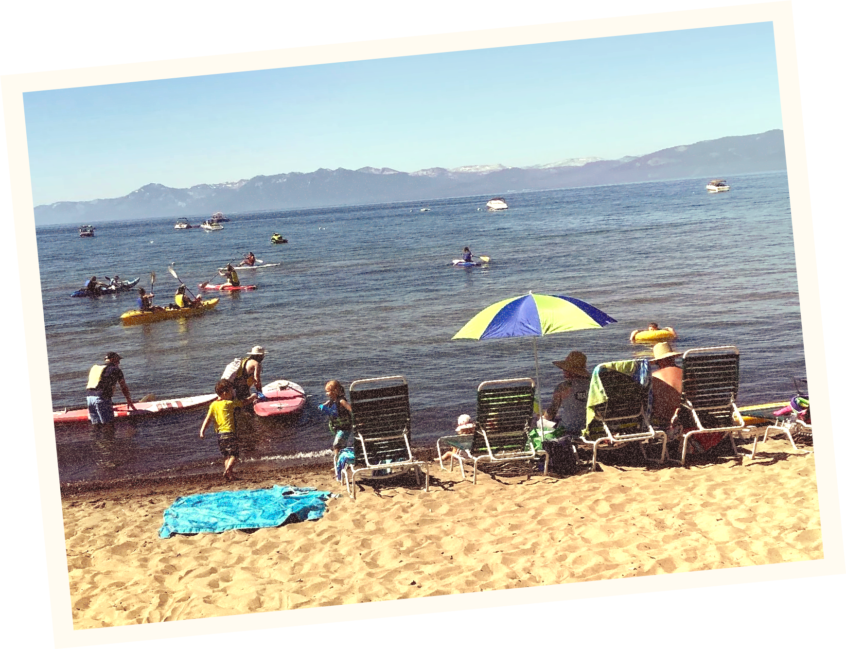 People enjoying a day at the beach with umbrellas, lounge chairs, and swimming, kayaking, and boating in the water, with mountains in the background.