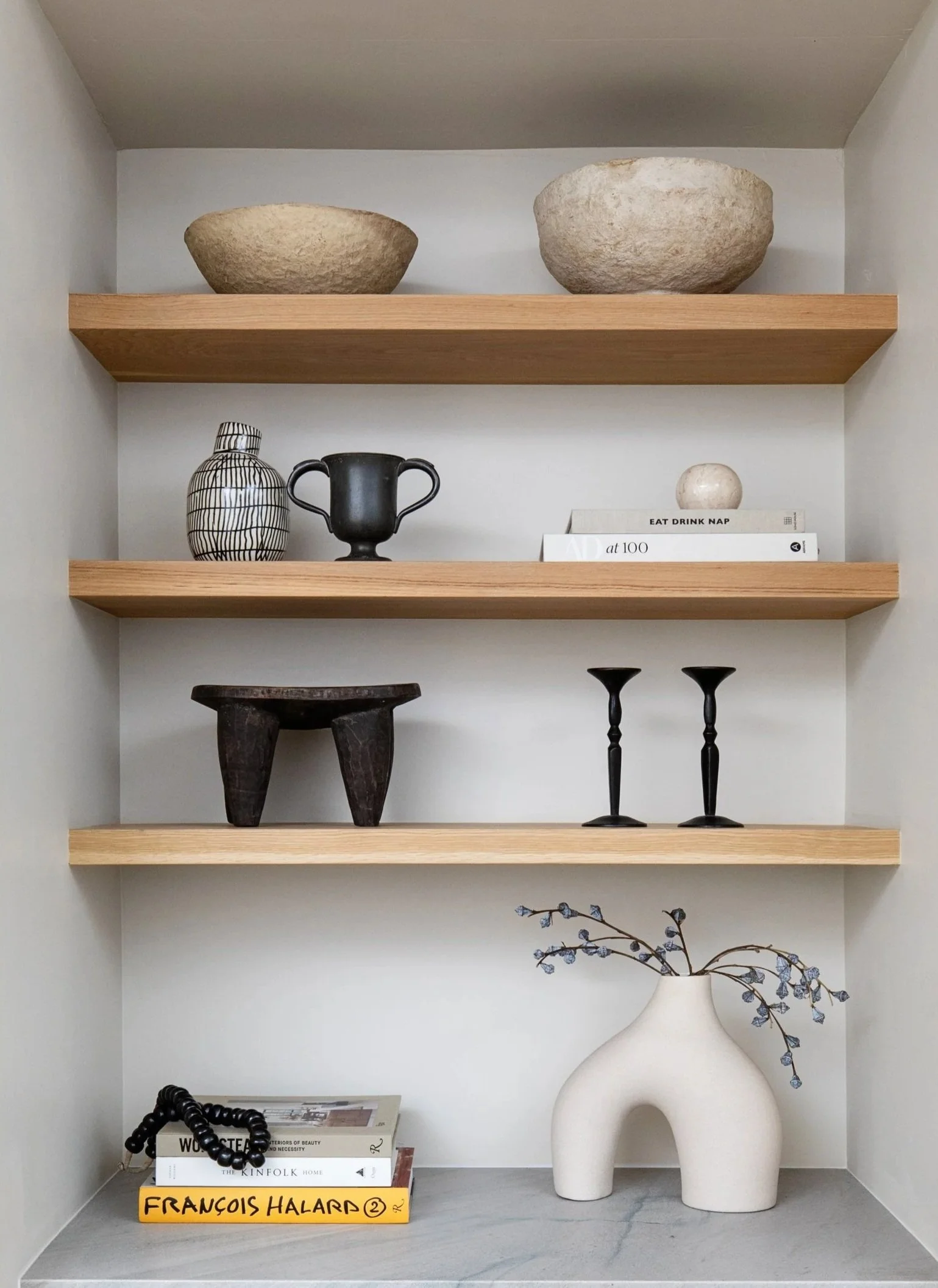 A built-in shelf with decorative items: two large stone bowls on the top shelf, a black and white patterned vase and a small black urn on the second shelf, a small dark wooden stool and two black candle holders on the third shelf, a white ceramic vase with blue flowers on the bottom shelf, and a stack of books along with a black beaded necklace on the marble surface in front.