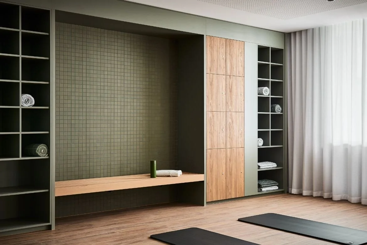 Wellness room or home yoga studio with custom built-in shelving, cabinetry and bench. A material palette of soft curtains, earthy green tile and paint, and wood flooring.