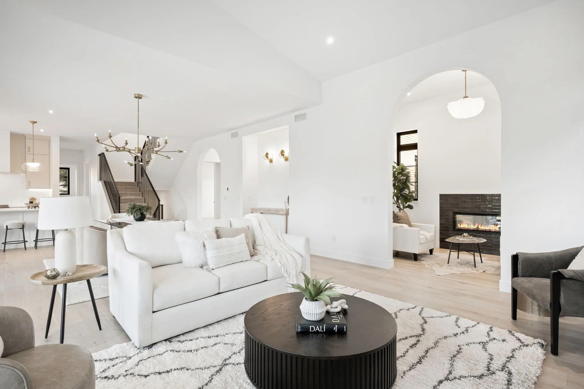 Bright living room with white sofa, round black coffee table with books and a plant, neutral-colored chairs, a fireplace, and natural light from windows.
