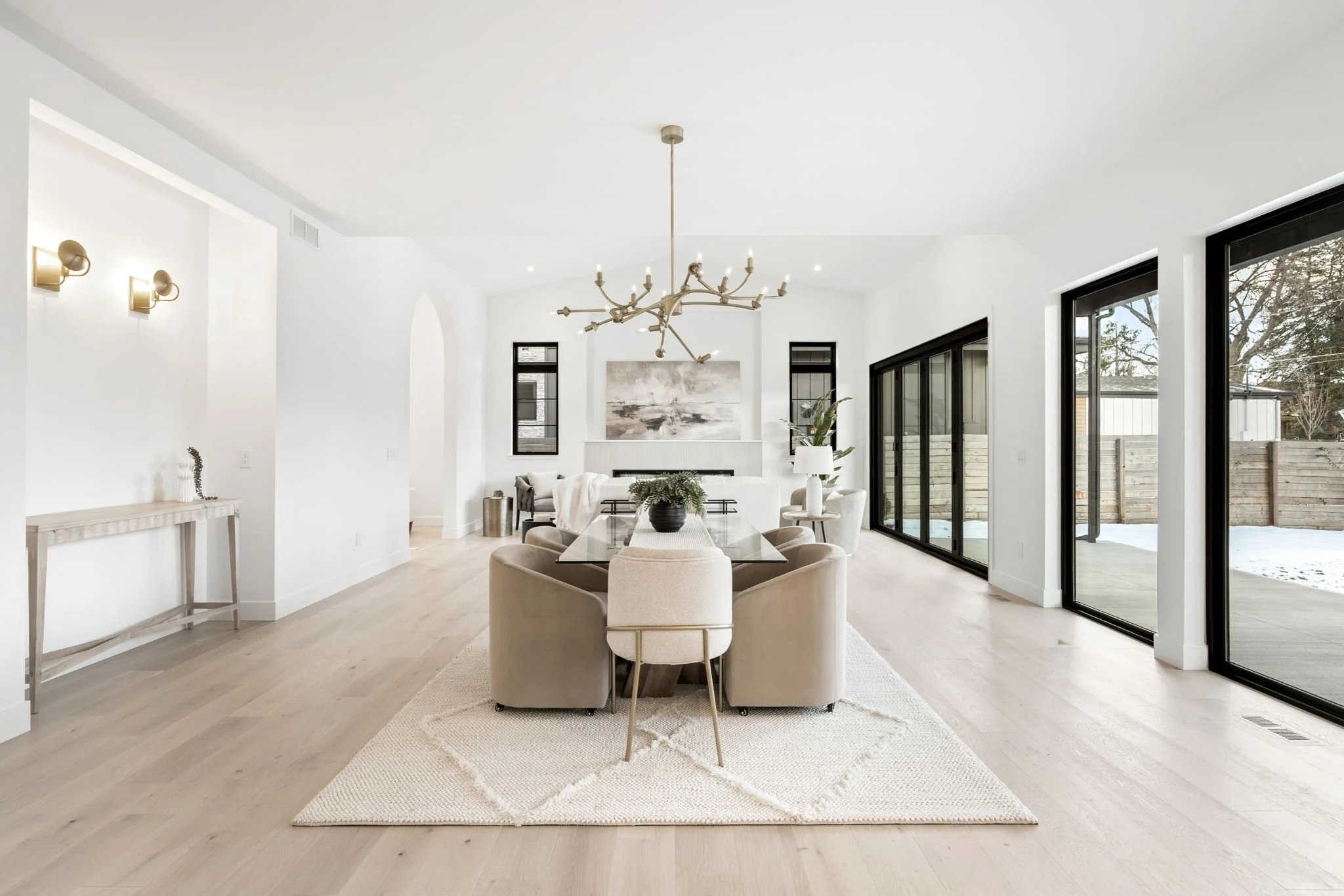 Bright open-concept living and dining room with light wood floors, white walls, modern chandelier, large glass sliding doors, and outdoor view.