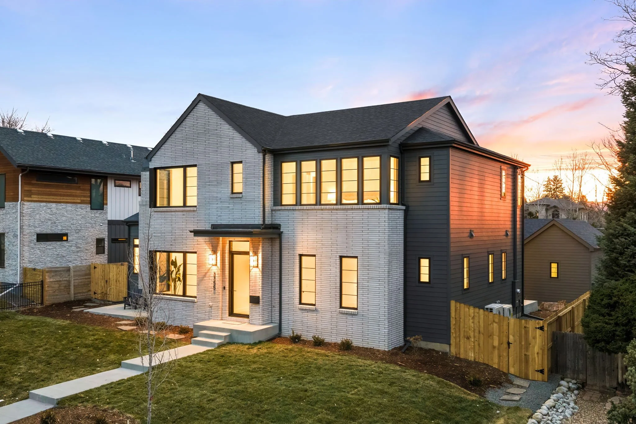 Modern two-story house with white brick and black siding exterior, illuminated windows, front steps, surrounded by a lawn and fenced yard, at sunset.