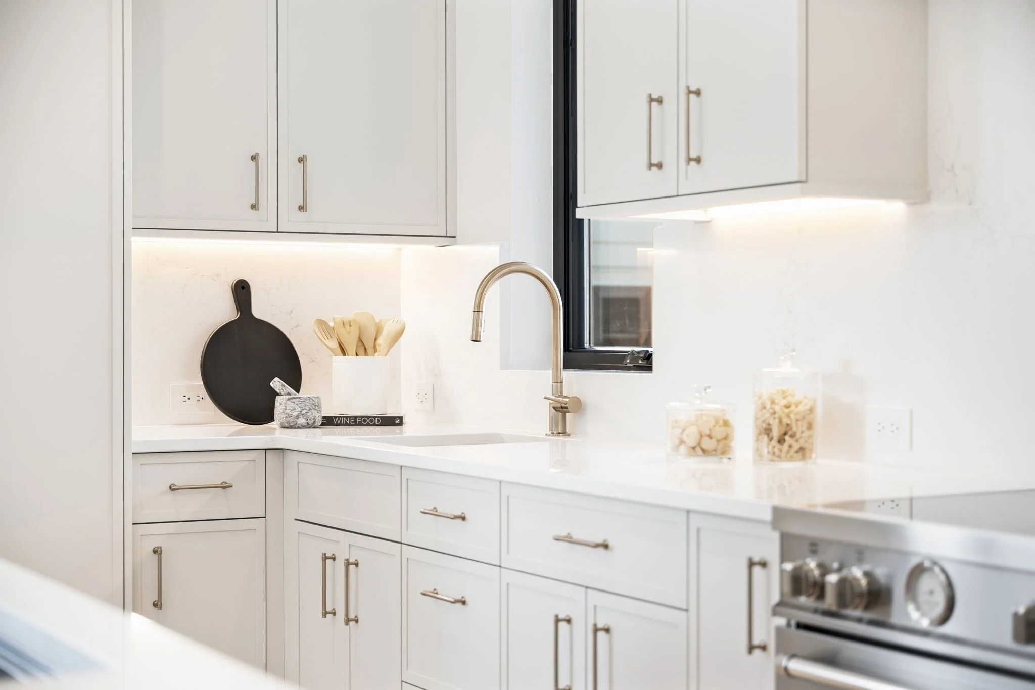 Modern white kitchen with black window frame, gold handles, countertop with jars and cutting board, and stainless steel stove.