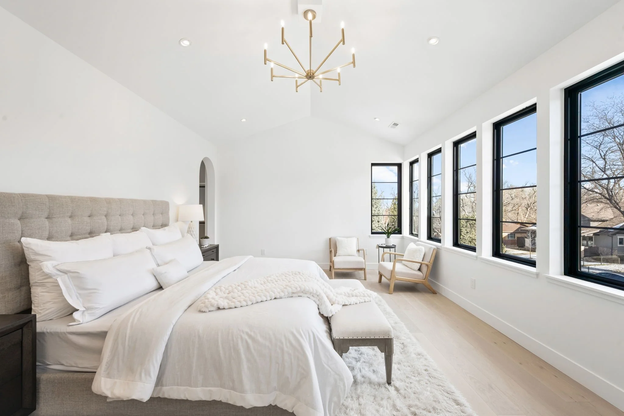 Bright, modern bedroom with a large bed, white bedding, beige headboard, small side tables, a black and gold chandelier, and multiple windows showing a sunny outdoor scene with trees.