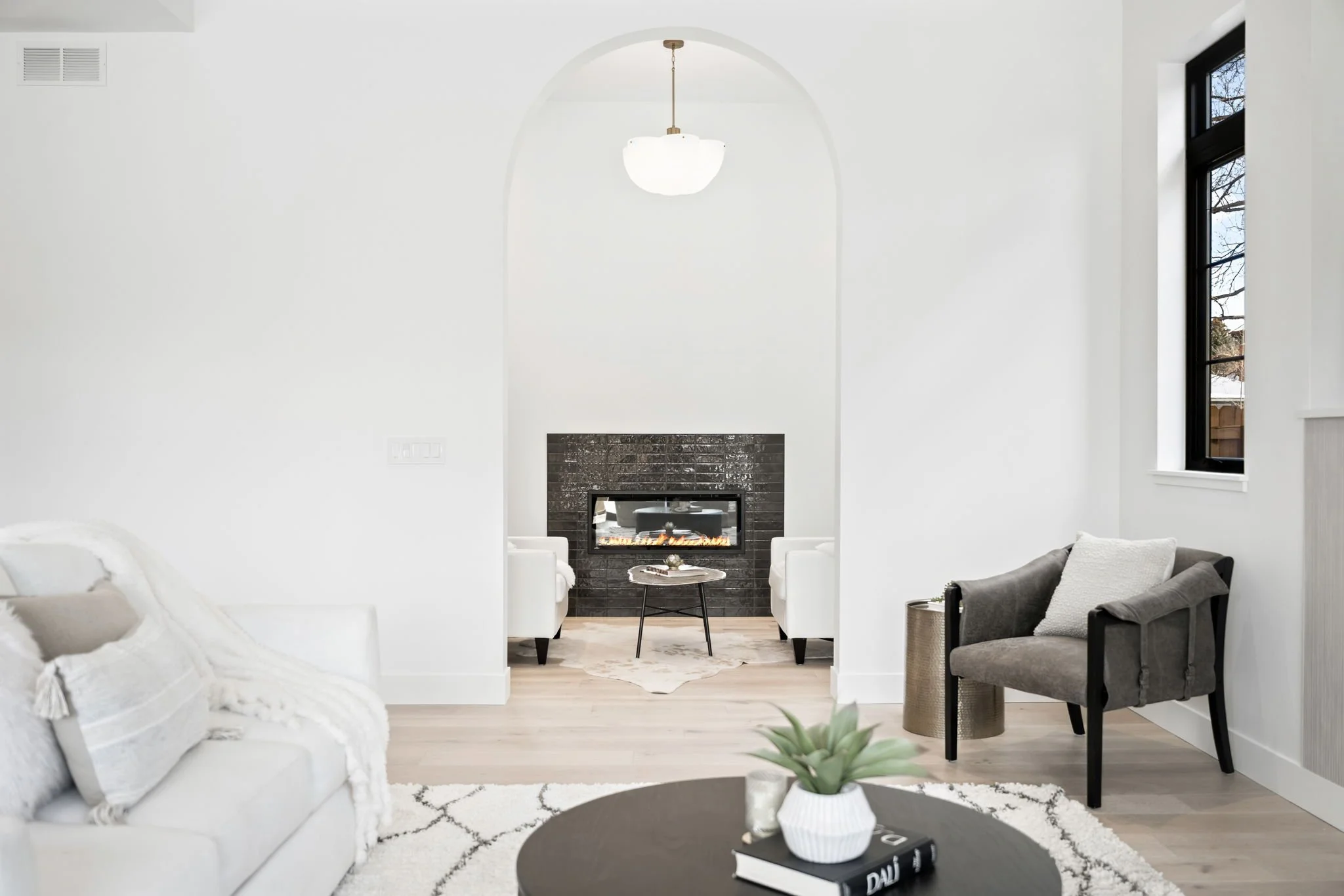 Modern living room with white walls, a ceiling light fixture, a fireplace with black tiles, a white armchair, a gray armchair, and a round black coffee table with decor items, including a plant, a book, and a candle.