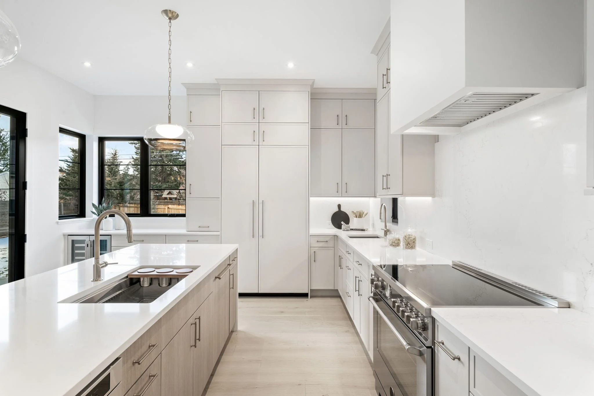Modern white kitchen with an island, stainless steel appliances, and large windows showing outdoor trees.
