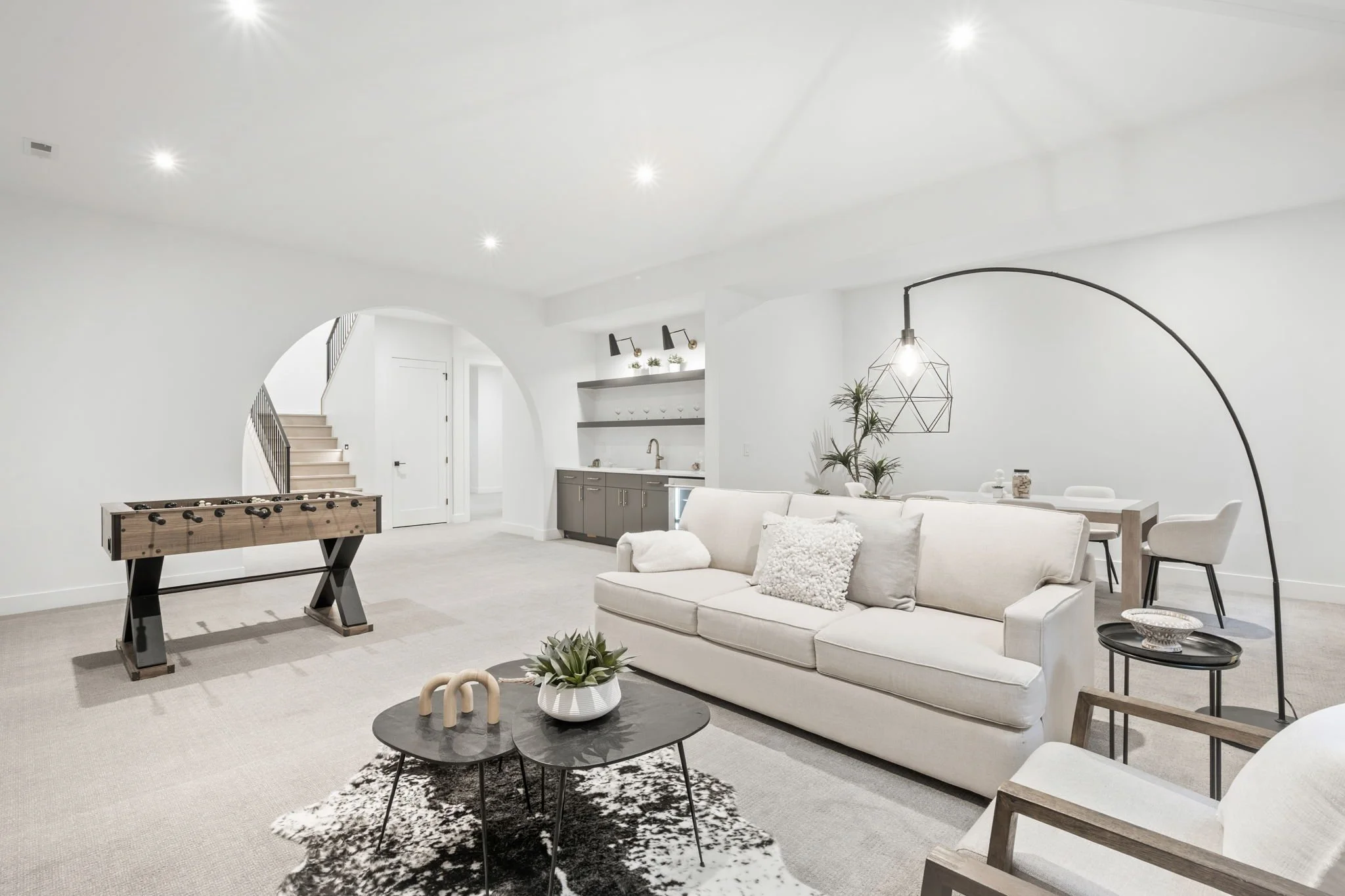 A spacious, modern living room with white walls, ceiling, and furniture, featuring a foosball table, a white sofa, and a black-and-white cowhide rug.