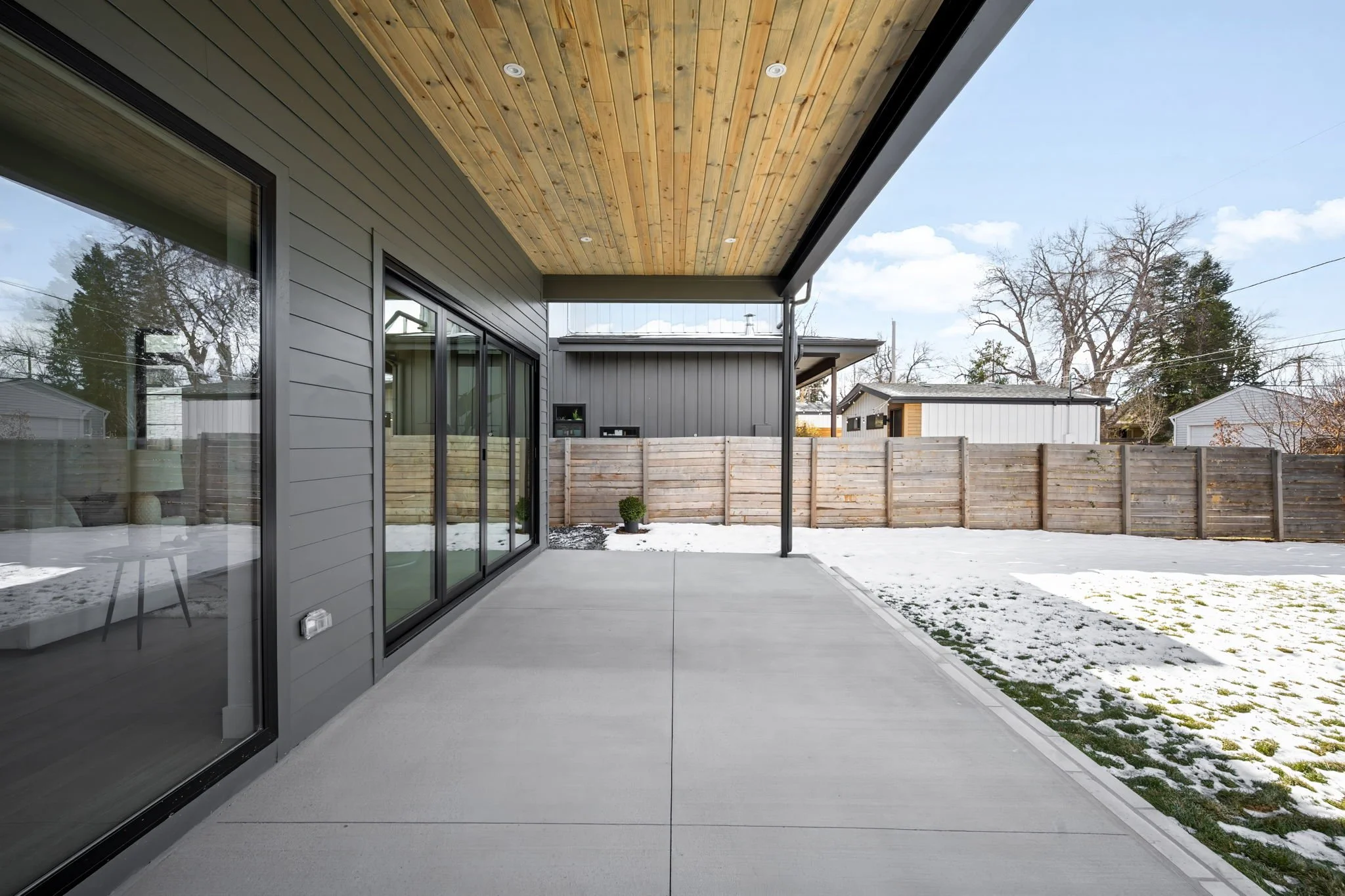 Modern backyard patio with gray concrete flooring, sliding glass doors, a wooden ceiling with recessed lighting, and a snow-dusted yard enclosed by a wooden fence.