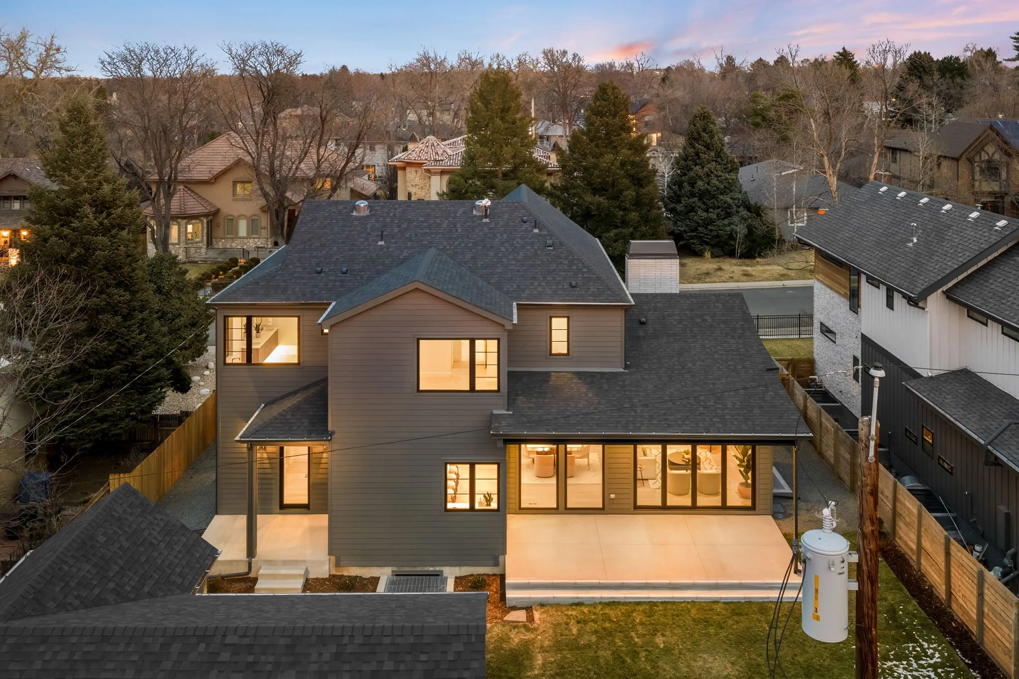 A modern two-story house in a suburban neighborhood at dusk, with warm interior lights on and a well-lit backyard patio.
