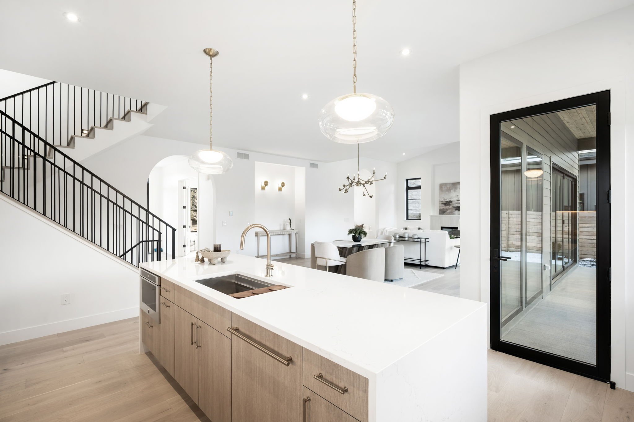 Modern open-concept kitchen and living area with white walls, wooden flooring, a large kitchen island, pendant lights, and glass doors leading to an outdoor space.