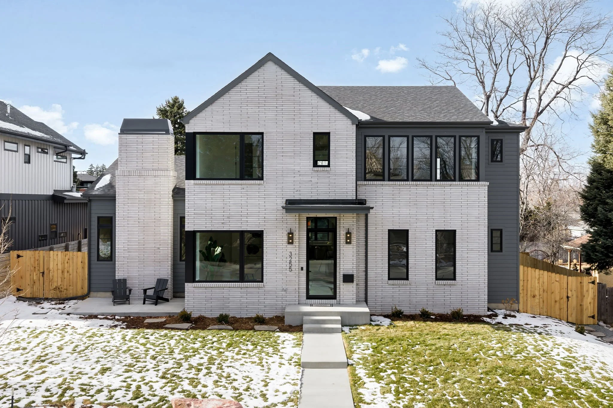 Modern two-story house with white brick and dark gray siding, black window frames, front porch with steps, and a small front yard with patchy snow and grass, during daytime with a clear sky.