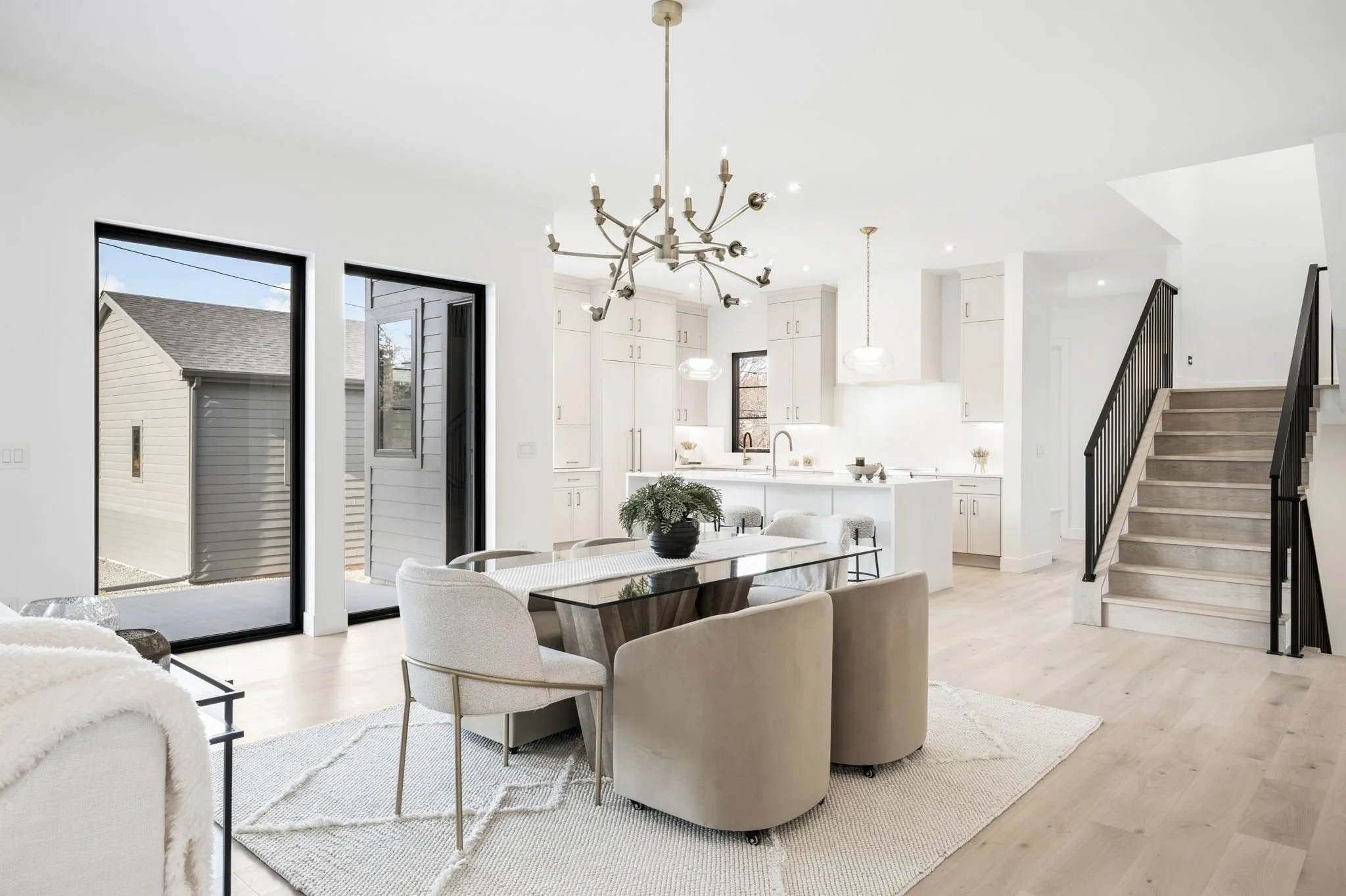 Bright, modern, open-concept kitchen and dining area with white cabinets, pendant lights, black-framed sliding glass doors, a glass-top dining table with beige and cream chairs, a staircase with black railing, and a light wood floor.