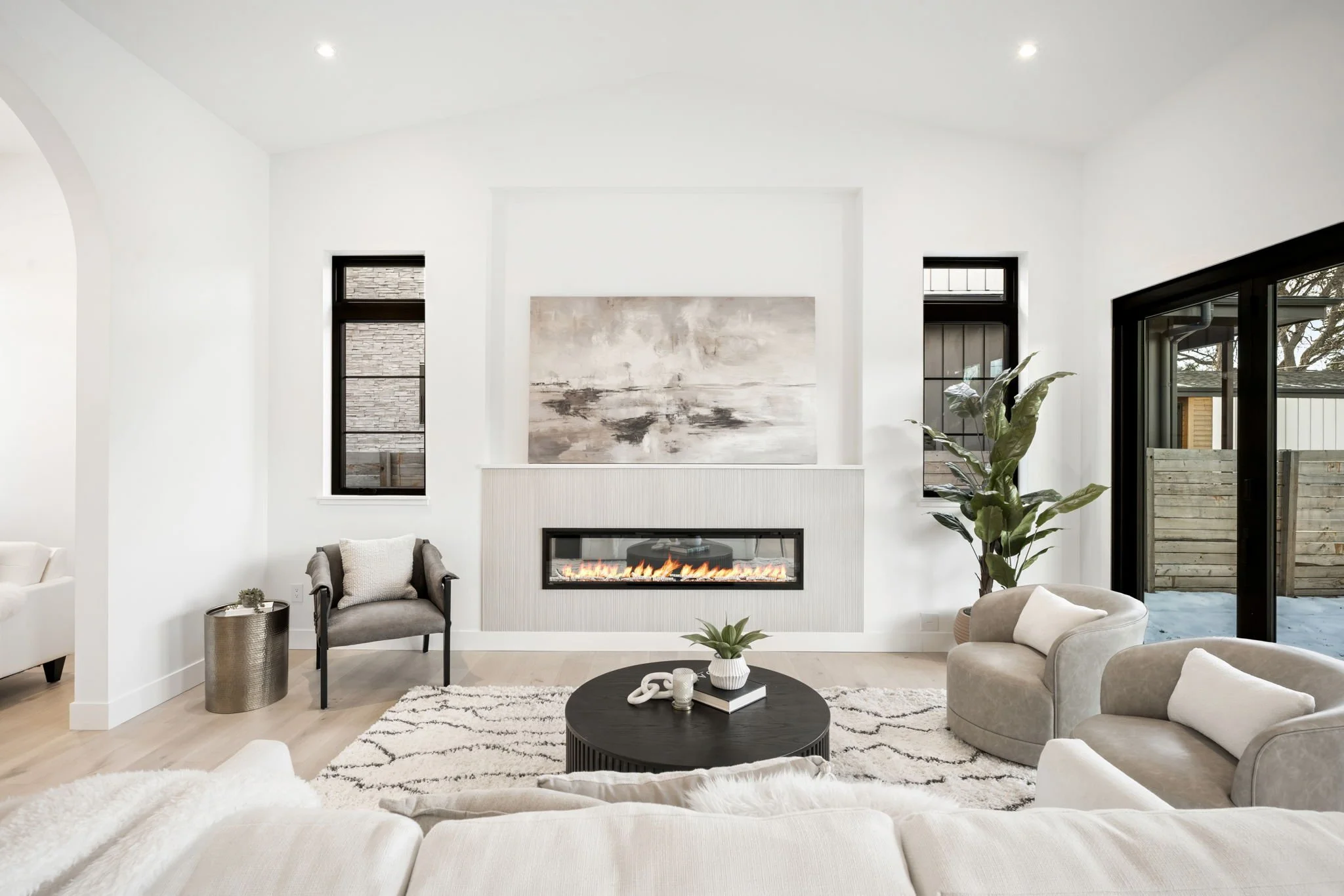 Modern living room with white walls, a fireplace, a black round coffee table, light-colored sofa and chairs, a large plant, and large window and sliding glass door leading to an outdoor space.
