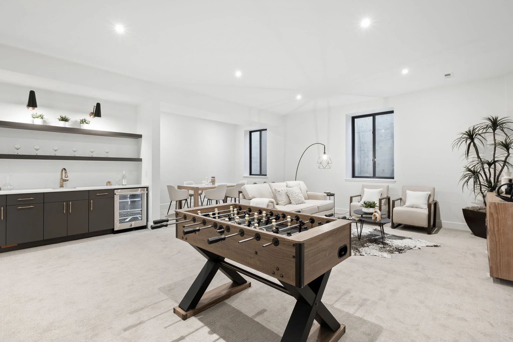 Modern living room with foosball table, white sofa, armchairs, dining table, kitchenette with dark cabinets, and potted plants.