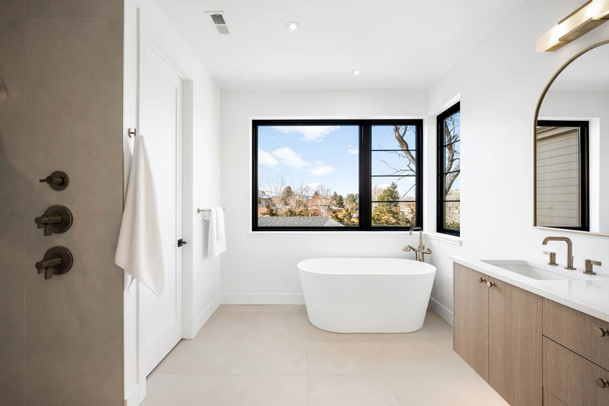 Modern bathroom with a white bathtub under large black-framed windows, a wooden vanity with a white countertop and gold fixtures, and a shower area with a white towel hanging.