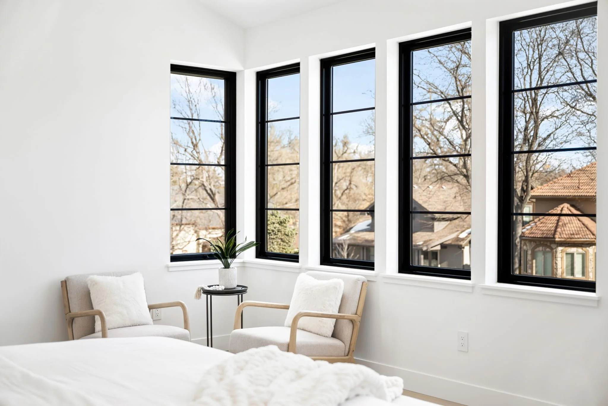 A bright, minimalist bedroom with large bay windows, white walls, and two beige armchairs with white pillows. A small black side table with a potted green plant is between the chairs.