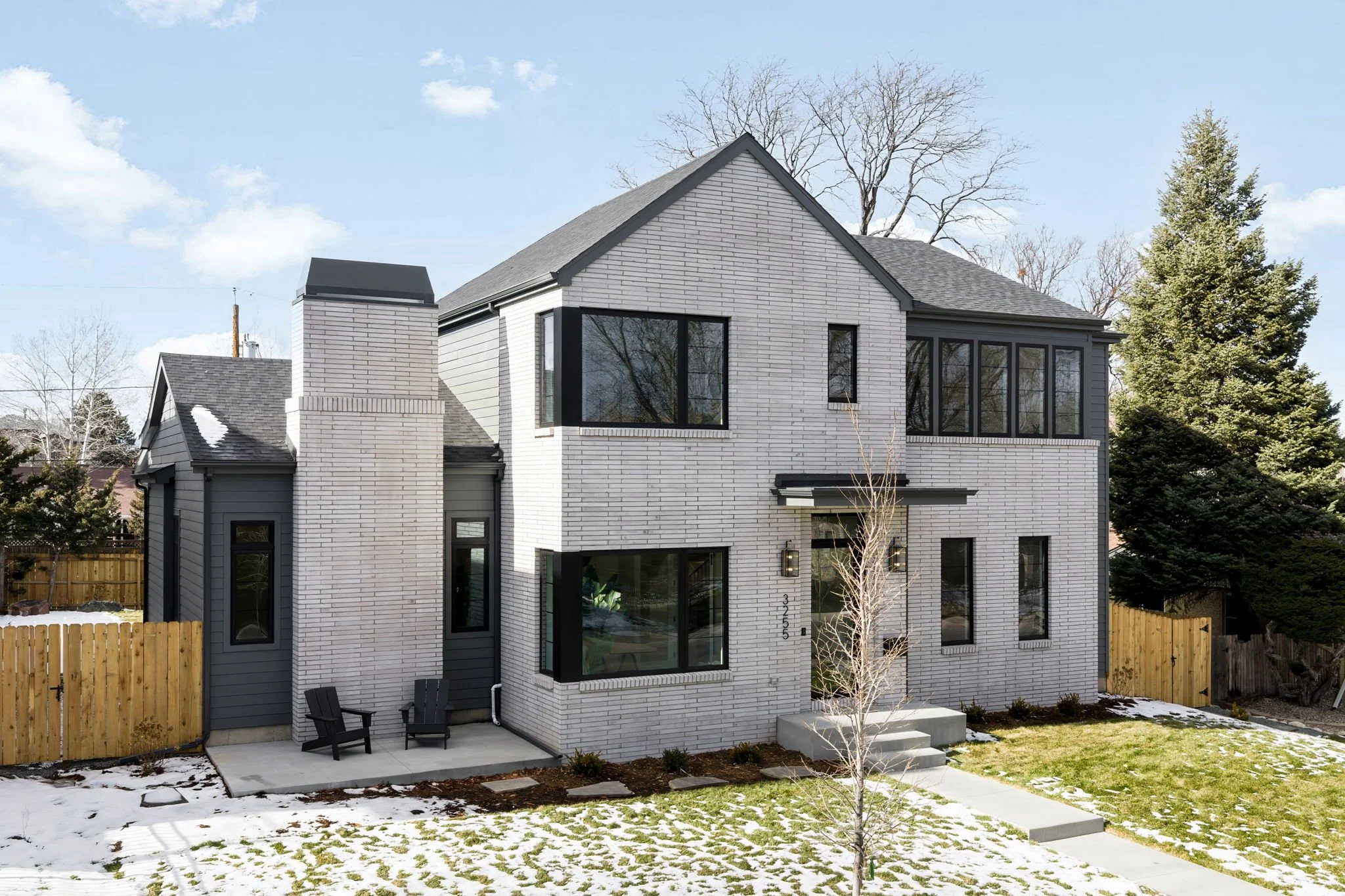 Modern two-story house with white brick exterior, black window frames, and a small front yard with snow and patches of grass, fenced with wooden panels.