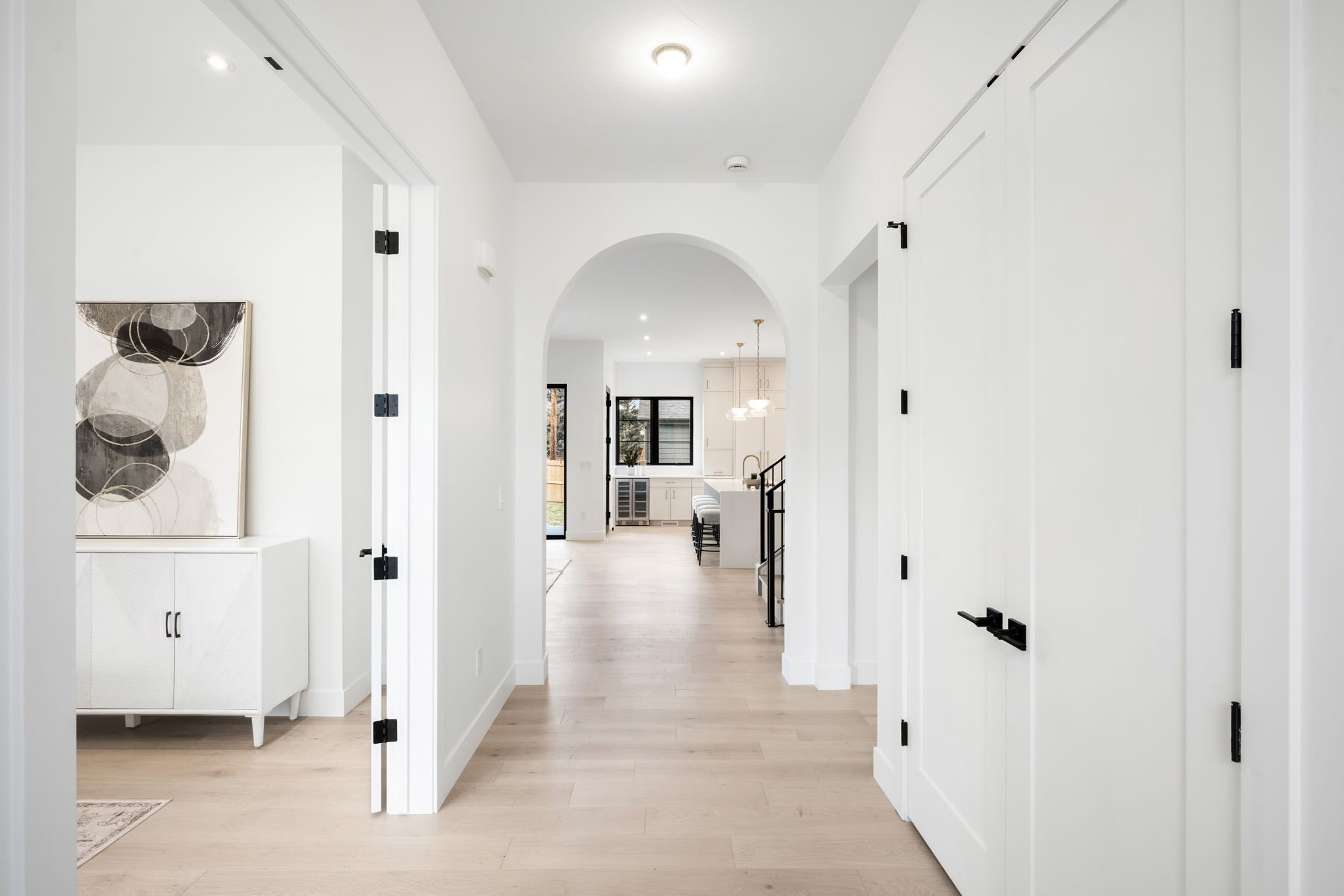 Interior view of a modern, minimalist home with white walls, light wood flooring, an arched doorway, and a kitchen area in the background with pendant lights and cabinetry.