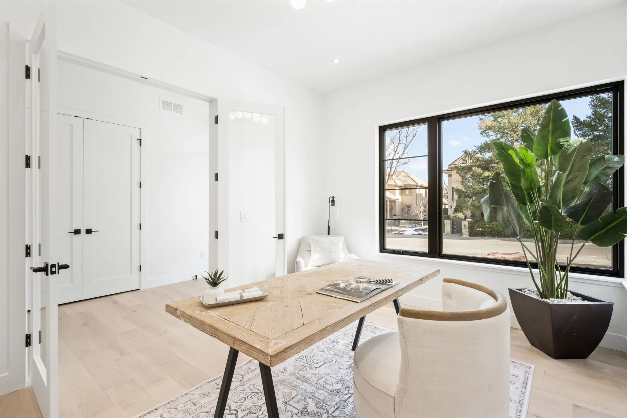 Bright, modern living space with white walls, a large window showing a suburban neighborhood, a wooden table, a white armchair, a black planter with large green leaves, and minimalist decor.