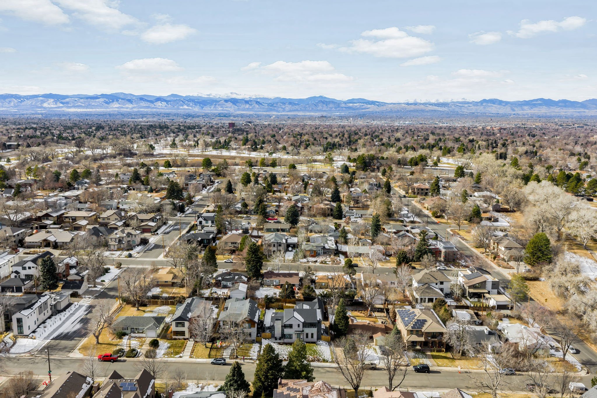 Aerial view of a suburban neighborhood with houses, trees, and streets, with snow patches, mountains in the distance, and a partly cloudy sky.