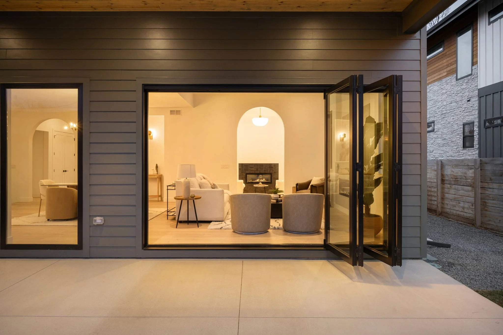 View of a modern house's open sliding glass door revealing a warmly lit interior living room with neutral-colored furniture, fireplace, and contemporary decor, outdoors a concrete patio with a wooden fence.