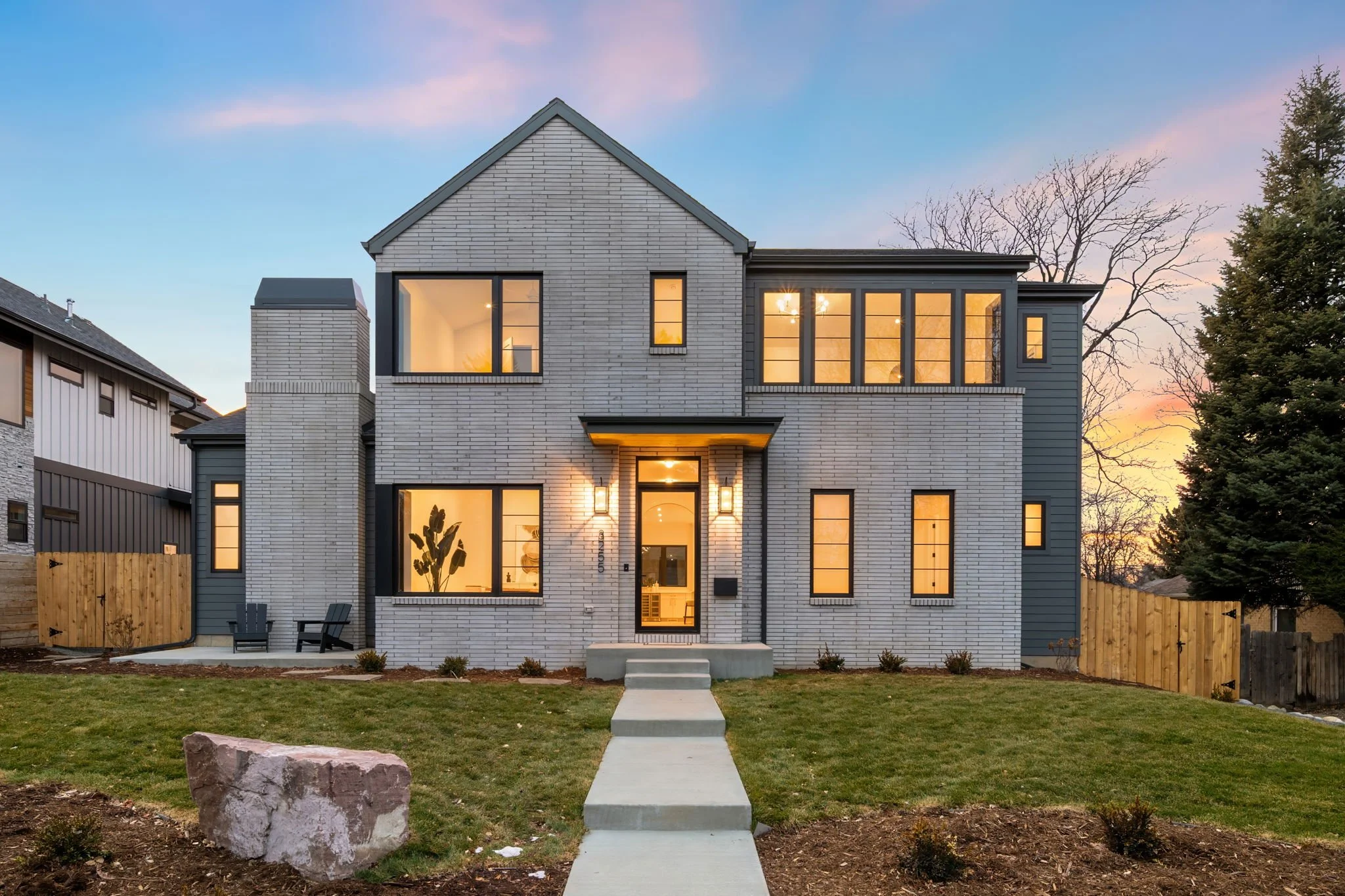 Modern two-story house with white brick and dark siding, front yard with grass, concrete walkway, and outdoor seating at sunset.
