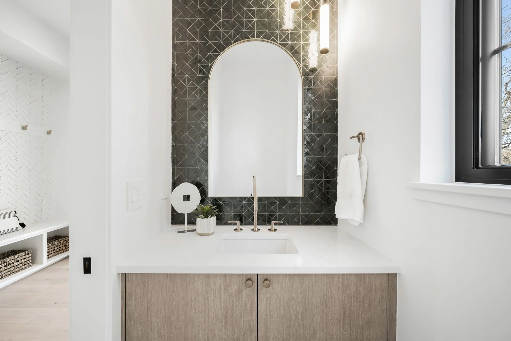 Modern bathroom vanity with a white countertop, a rectangular sink, and a black geometric tiled wall. A large arched mirror is centered behind the sink, with hanging light fixtures above. A small potted plant and a decorative circular mirror sit on t