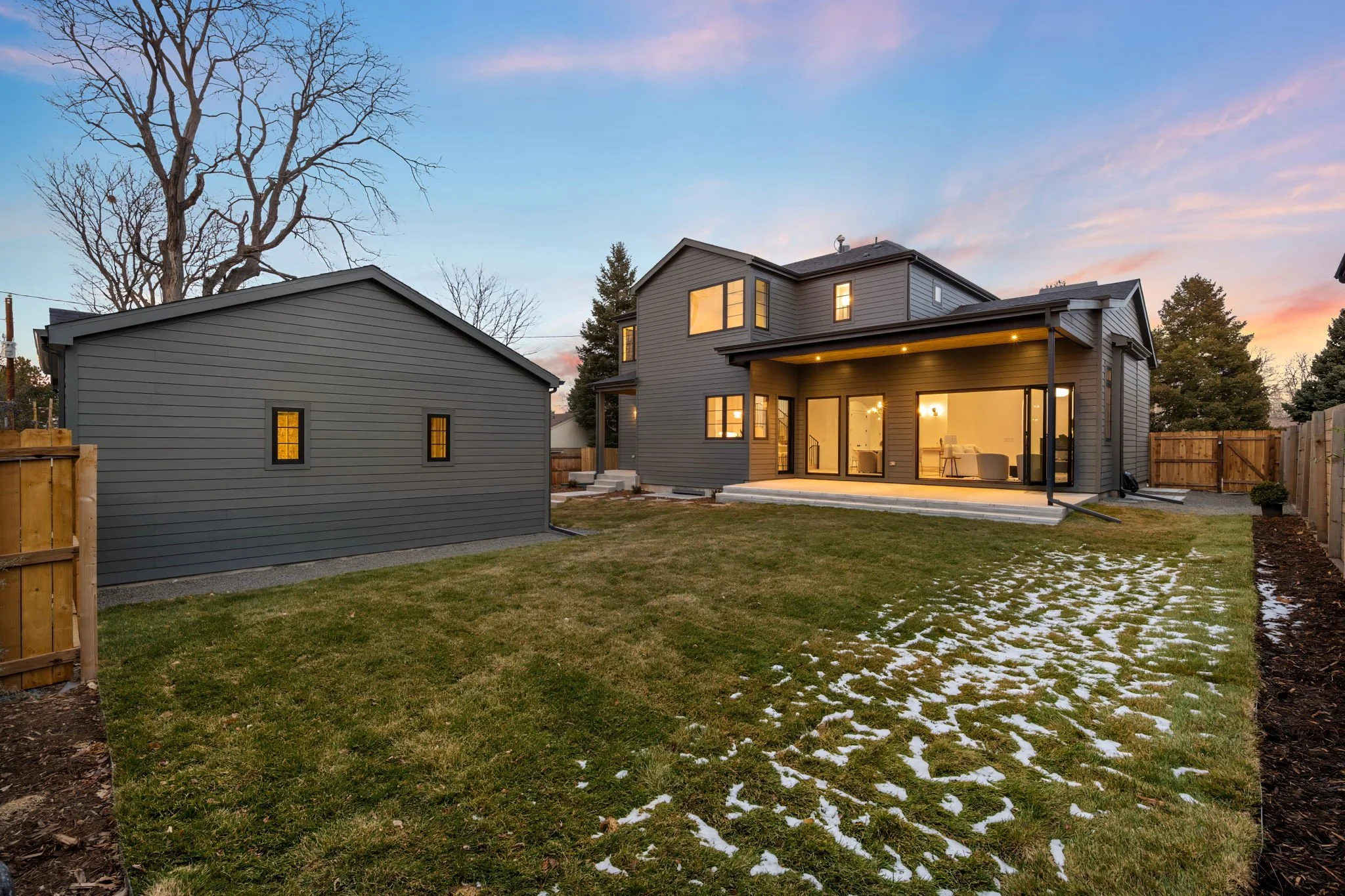 Modern gray house with large glass doors, illuminated interior, and a small detached gray shed in the backyard during dusk. There is some snow on the grass and a wooden fence surrounding the yard.