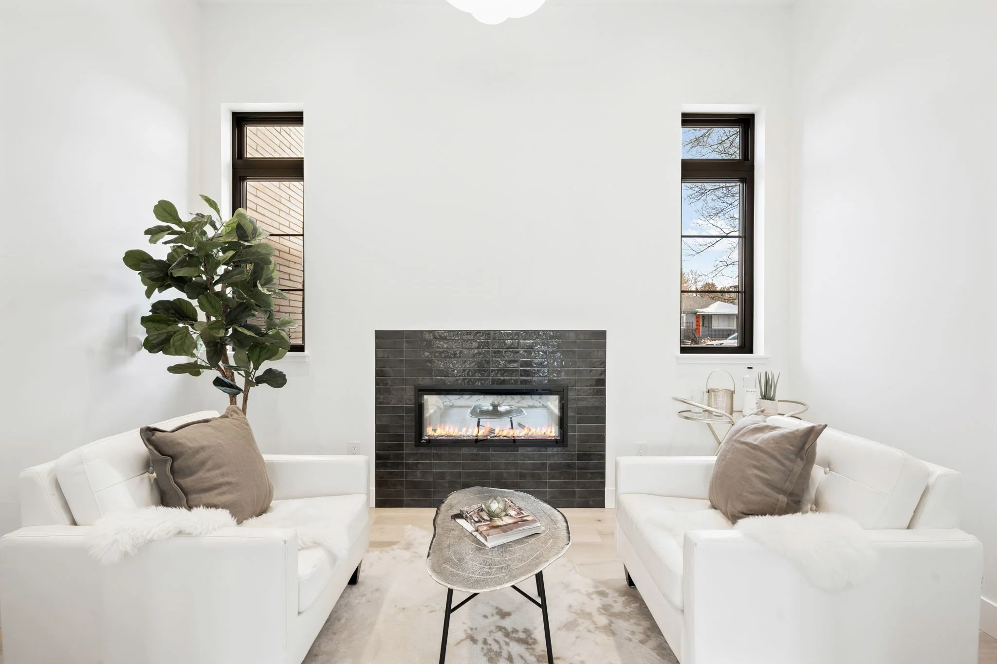 A modern living room with white sofas, a black fireplace, and two tall windows showing a neighborhood outside. Decor includes a large potted ficus, cushions, a small side table, and a coffee table with magazines.