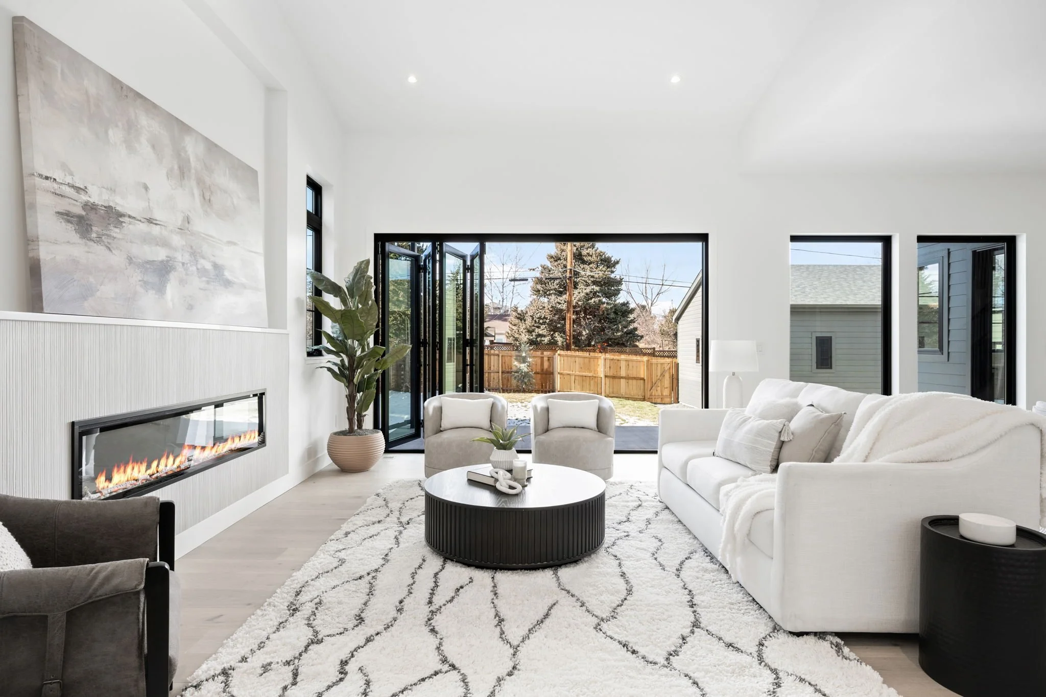 Modern living room with white sofa, beige armchairs, black round coffee table, white rug, fireplace, large windows, and sliding glass doors leading to backyard.