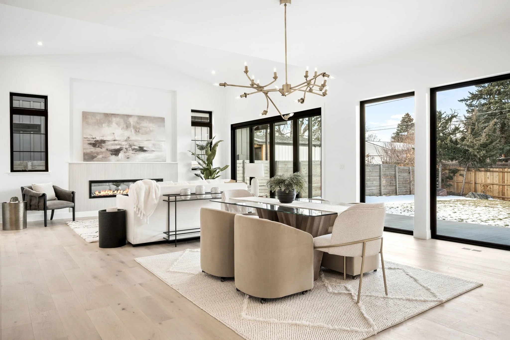 Modern living room with white walls, large sliding glass doors, and a light wood floor. Features include a white sofa with a blanket, a black armchair, a fireplace, a dining table with chairs, a chandelier, and outdoor view with snow and trees.