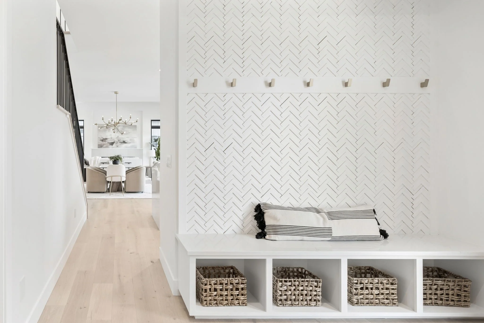 Interior view of a modern minimalist white entryway with built-in bench, woven baskets, and decorative wall hooks, leading to a dining area with a chandelier and round table.