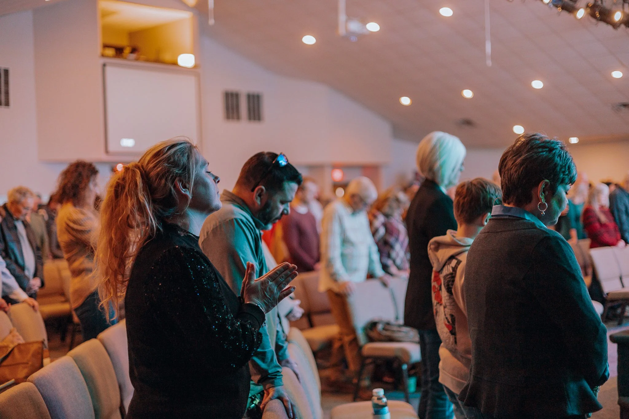 People standing in prayer in church with eyes closed and hands raised or clasped.