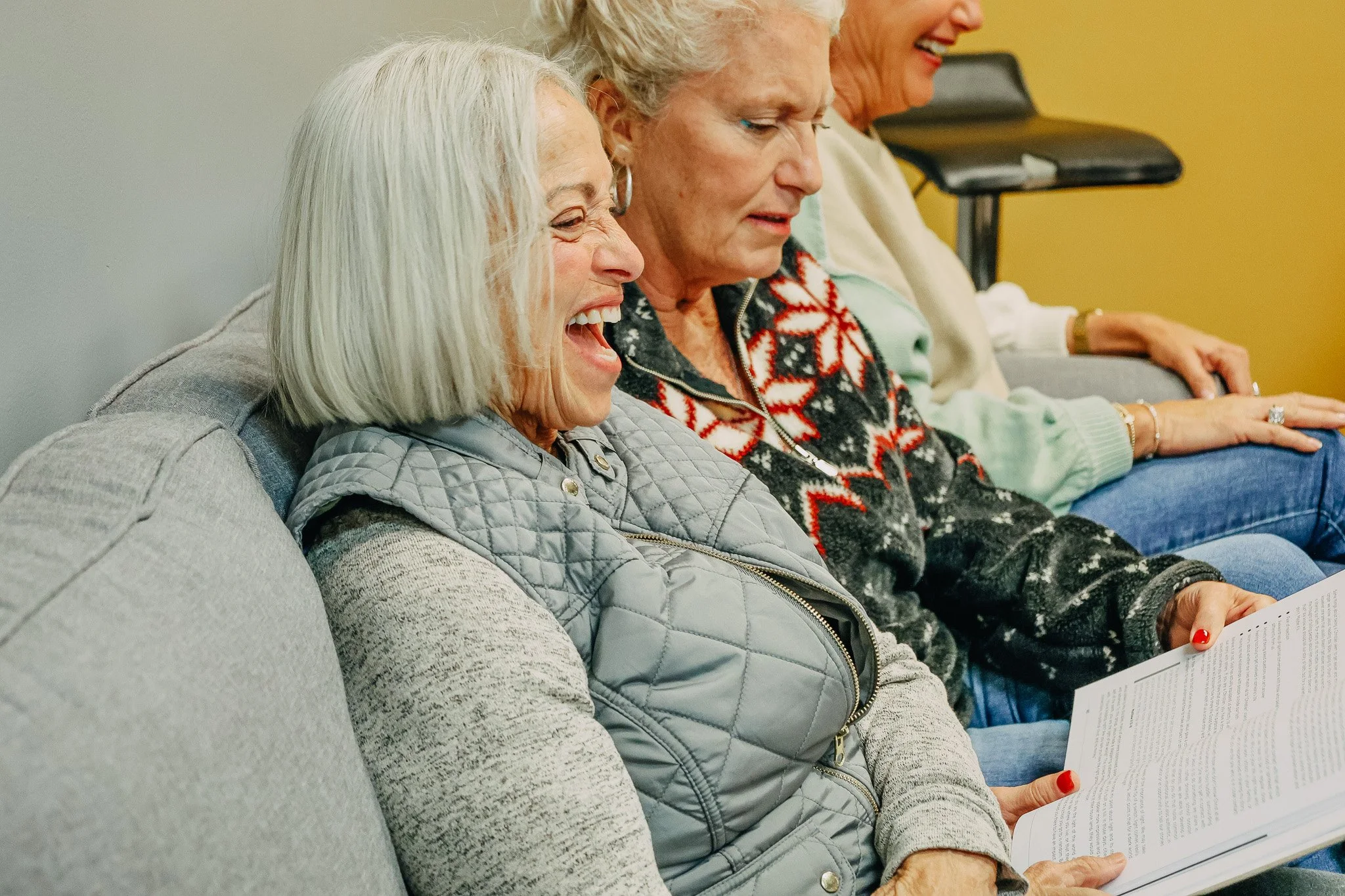 Three women sitting on a couch, laughing and reading a book together at a life group.