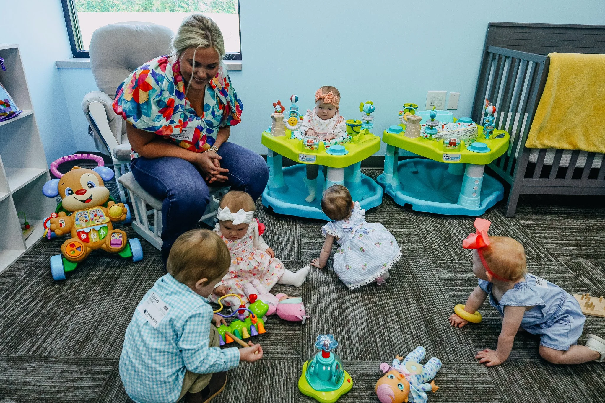 A woman and five young children in a playroom with toys, including baby activity centers, stuffed animals, and a small carousel, on a carpeted floor near a crib and a window.