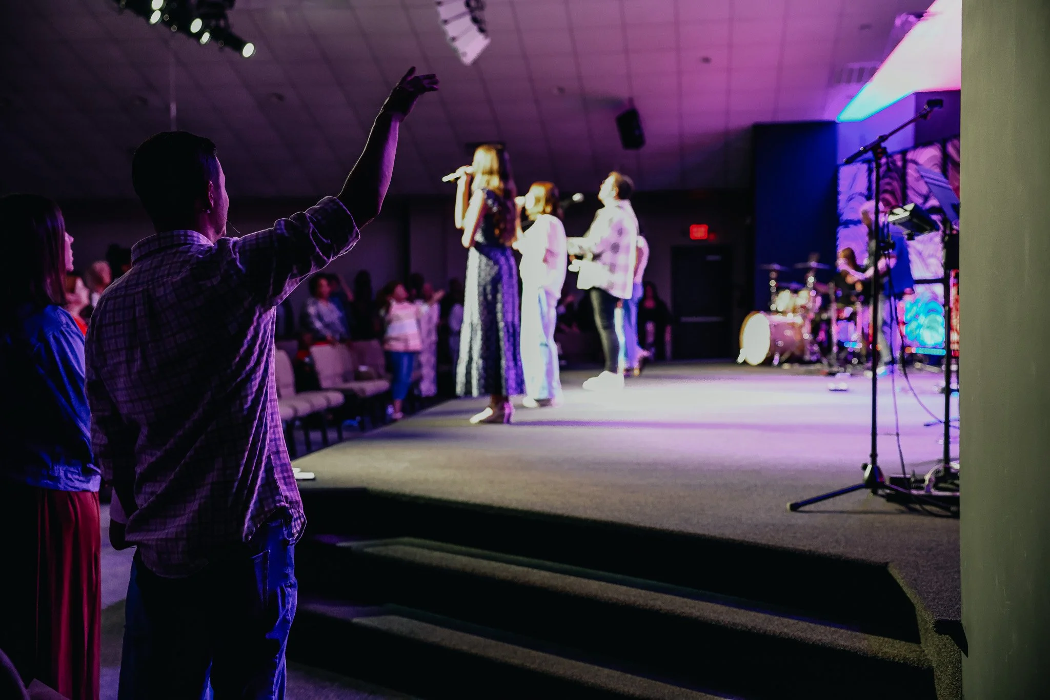 People worshipping on a stage with a musician, backed by stage lights and musical instruments, in a worship service.