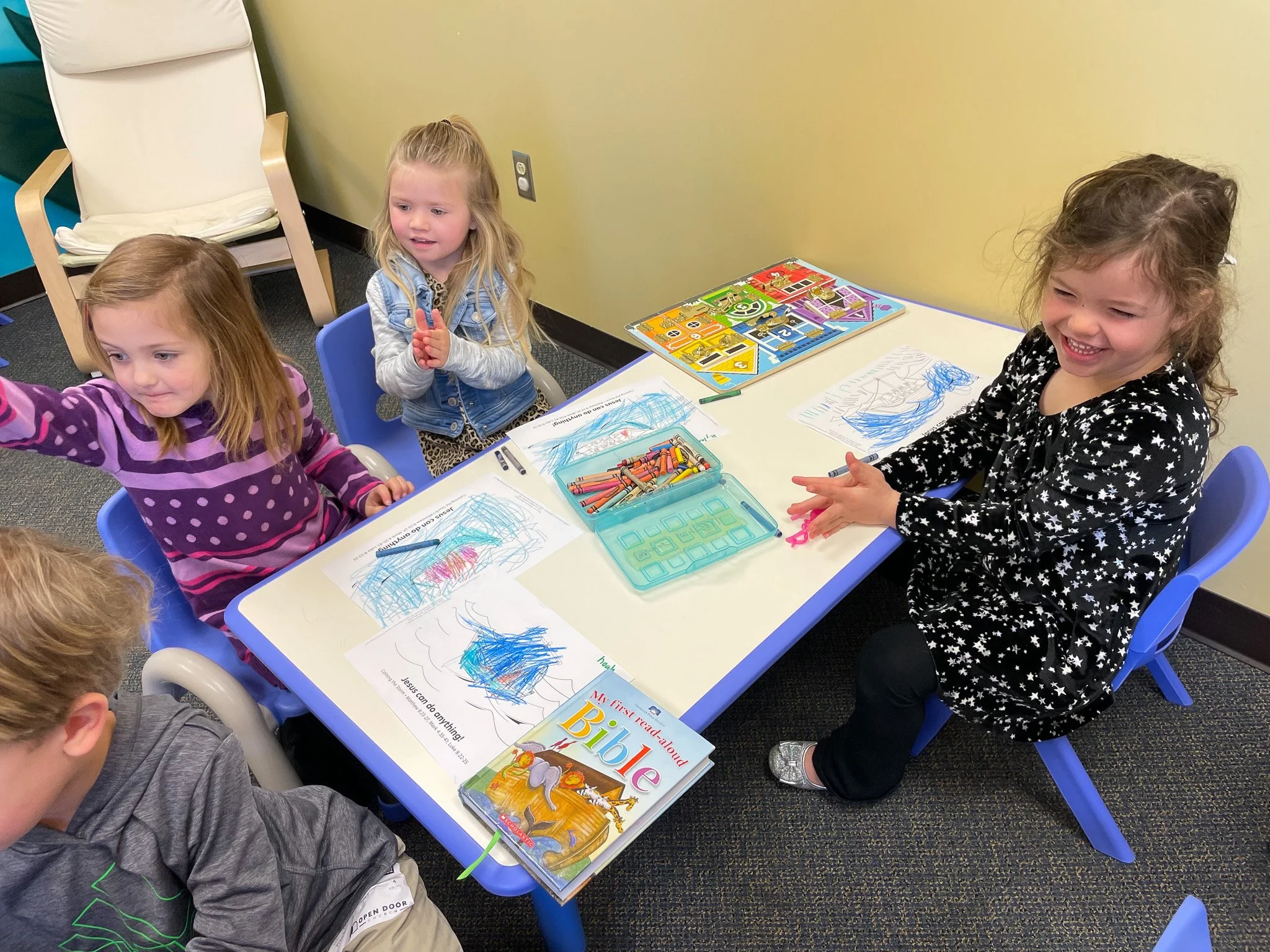 Four young girls sitting at a table in a classroom, engaged in drawing and coloring activities. The table has crayons, coloring pages, and a children's Bible. The girls are smiling and appear to be enjoying the activity.