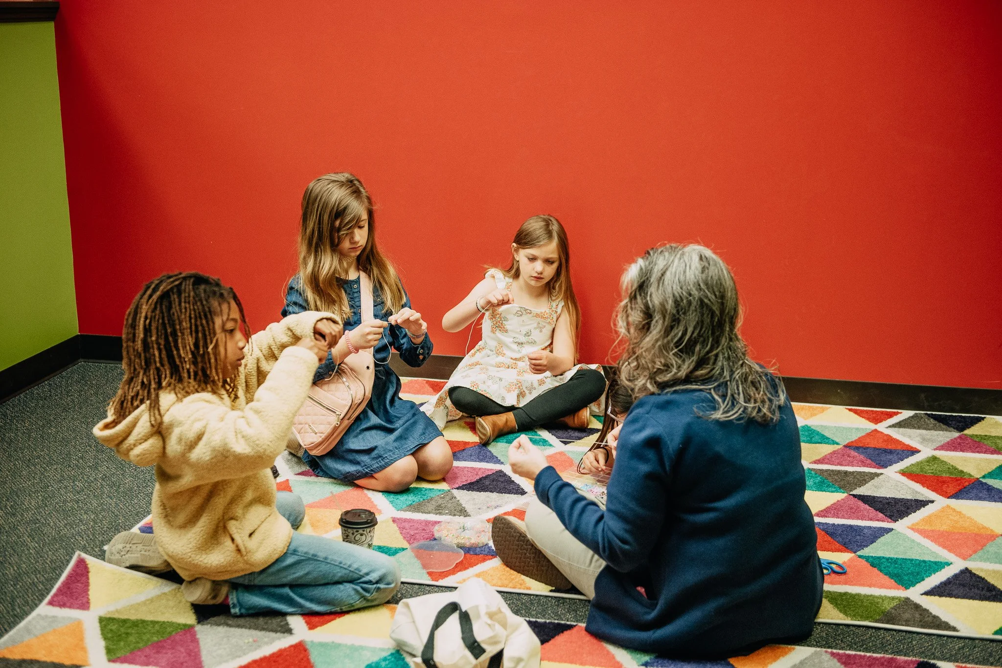 Four children sitting on a colorful patterned carpet in front of a red wall, engaged in a craft activity with a leader at Open Door Kidz.
