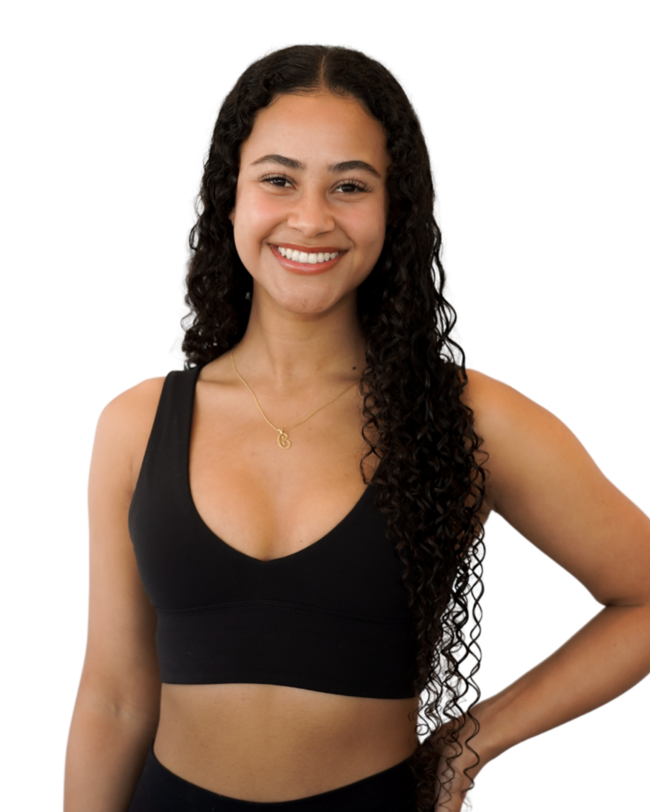 A young woman with long curly hair, smiling, wearing a black sports bra and black leggings, and a gold necklace with a pendant.