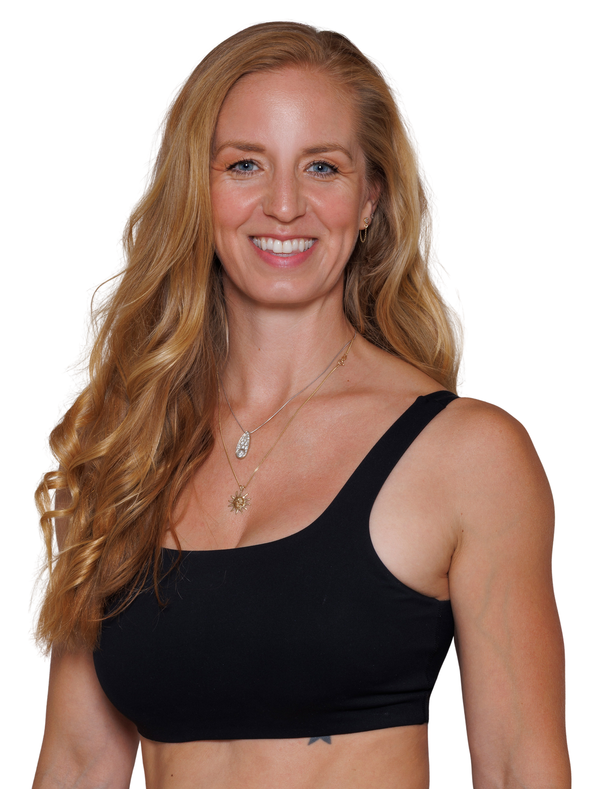 The woman with long strawberry blonde curled hair, wearing a black sports bra and two necklaces, one gold necklace of a sun and another silver elongated necklace, smiling at the camera.