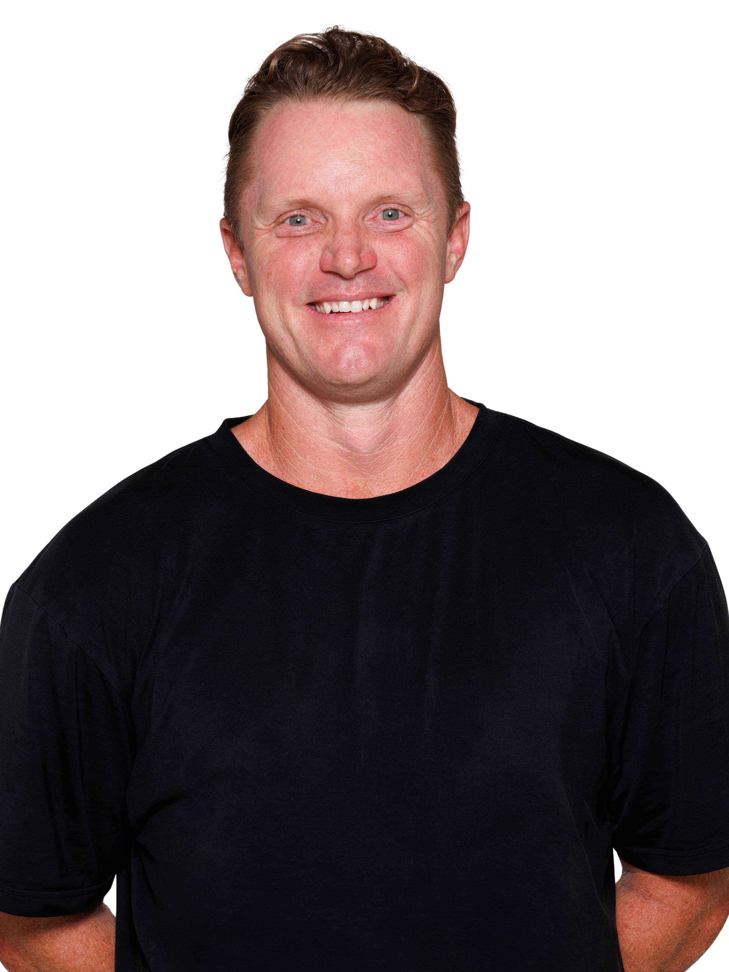 A smiling man with short, light brown hair, wearing a black T-shirt, standing with arms behind back against a plain white background.