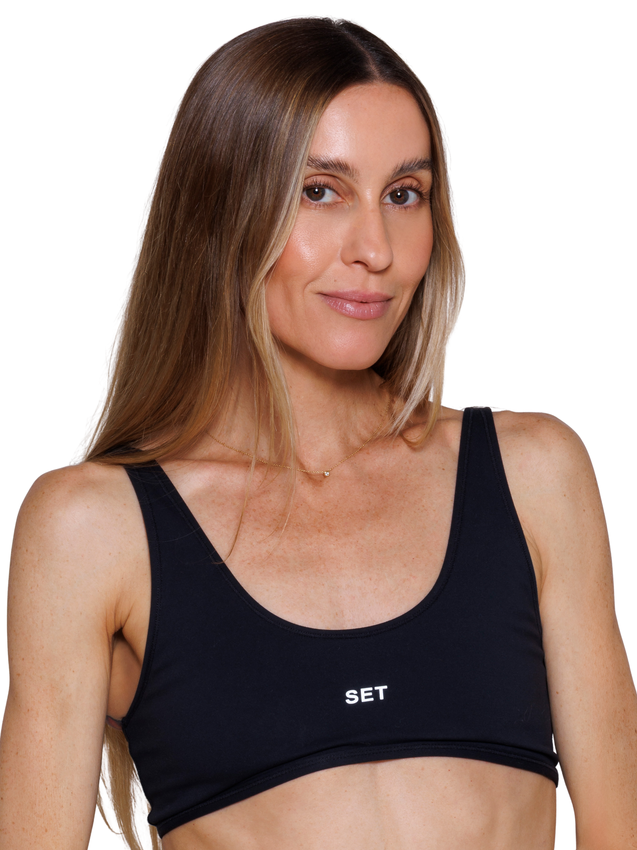 A woman wearing a black sports bra and gold jewelry, smiling and posing with hands on hips against a white background.