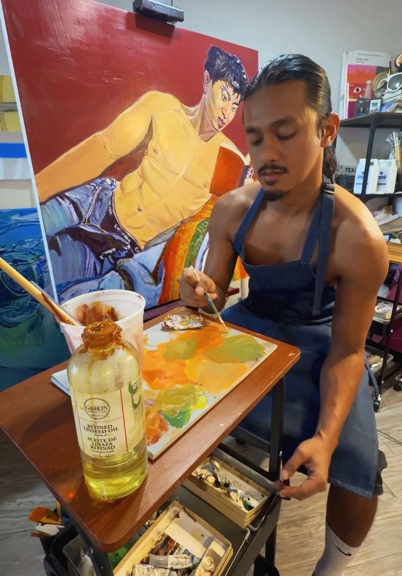 An artist is painting a portrait of a shirtless man in a painting studio, with art supplies and a partially completed colorful artwork in the background.