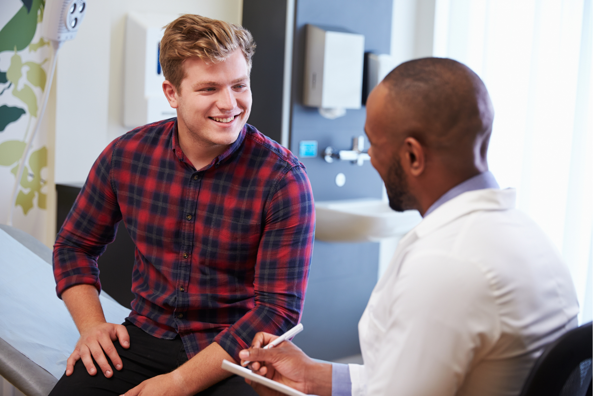 A study participant sits on an exam table, smiling as he converses with a doctor. The doctor is filling out a form.