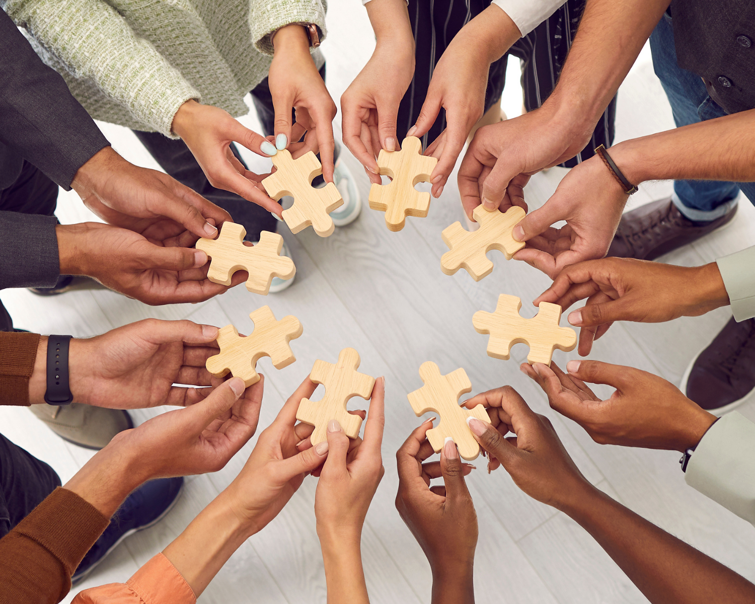 Multiple people of various ethnicities stand in a circle, holding out large wooden puzzle pieces towards the center as they each contribute a piece to the puzzle.