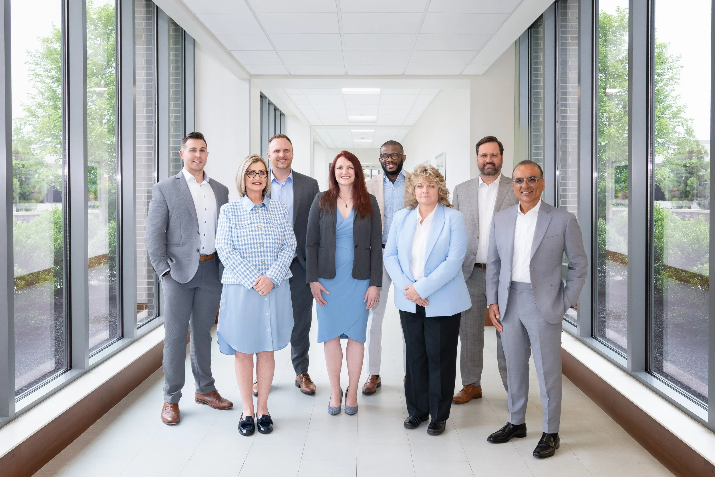 Group of eight diverse professionals standing in a bright office corridor with large windows and outside greenery.