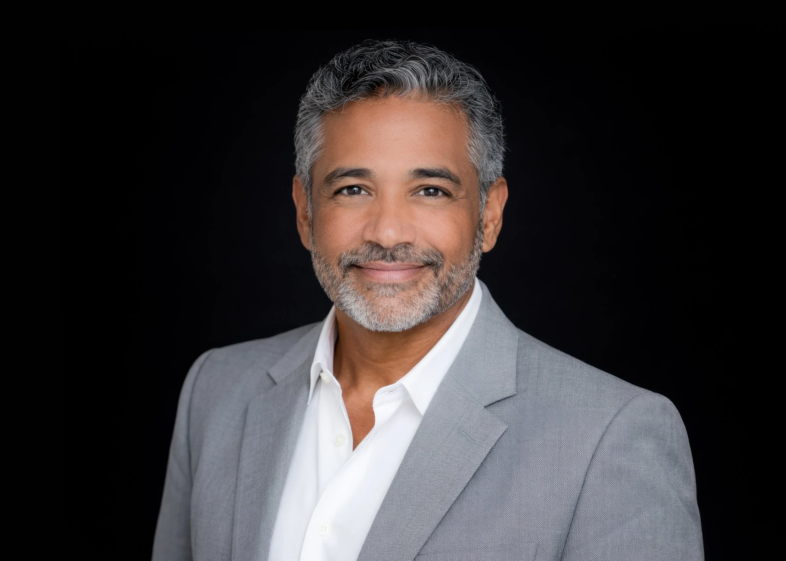 Headshot of a man with gray hair and a beard, wearing a light gray suit and white shirt, smiling against a black background.
